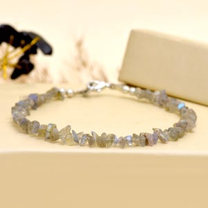 Könnte beinhalten: Ein zartes Armband mit kleinen, unregelmäßig geformten, hellgrauen Labradoritsteinen. Die Steine sind mit einer silberfarbenen Kette und einem Verschluss aufgereiht. Das Armband wird auf einer neutral getönten Oberfläche präsentiert.