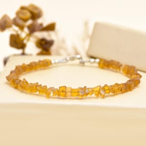 Könnte beinhalten: Ein zartes Armband mit kleinen, unregelmäßig geformten, bernsteinfarbenen Edelsteinen, die aneinandergereiht sind. Das Armband wird mit einem silberfarbenen Verschluss befestigt. Die Edelsteine haben ein facettiertes Aussehen und fangen das Licht ein. Das Armband wird vor einem neutralen Hintergrund präsentiert.