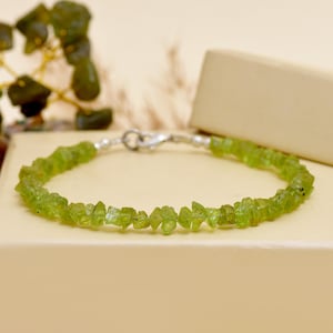 Puede incluir: Una delicada pulsera con piedras de peridoto verde claro en bruto, ensartadas en una cadena de plata. La pulsera se exhibe sobre una superficie de tonos neutros, resaltando la belleza natural de las piedras.