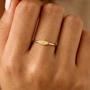 May include: A thin gold band ring featuring a small, horizontal, oval-shaped plaque at the centre. The plaque is engraved with the uppercase letter J. The ring has a polished, metallic finish and a simple, slender design that encircles the finger.
