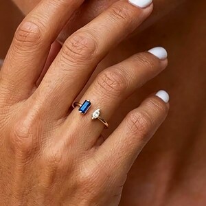 Op de afbeelding: Een open gouden ring met een rechthoekige blauwe saffier in baguette-slijpvorm aan het ene uiteinde en een heldere witte diamant in marquise-slijpvorm aan het andere. De ringband is dun en gepolijst met een opening tussen de stenen.