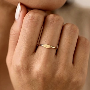 Puede incluir: Un anillo fino de oro que presenta una pequeña placa plana de forma ovalada horizontal en el centro. La placa está grabada con la letra minúscula j. La banda es lisa y circular, manteniendo un grosor constante alrededor del dedo.