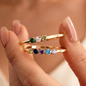 Puede incluir: Dos anillos de oro con piedras preciosas de corte redondo. El anillo superior tiene tres piedras en verde, blanco y verde claro. El anillo inferior tiene cinco piedras en amarillo, azul oscuro, morado, azul claro y rosa claro en una banda de oro.