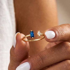 Peut inclure: Une bague en or avec un design à tige fendue. La bague présente une pierre précieuse rectangulaire bleu foncé et une petite pierre précieuse claire de forme ovale. La bague est tenue par une personne.