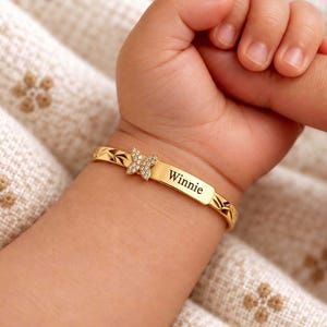 Könnte beinhalten: Ein goldfarbener Armreif mit einem kleinen, mit klaren Steinen besetzten Schmetterlingsanhänger und einer rechteckigen Namensplatte, in die der Name Winnie eingraviert ist. Das Band hat ein texturiertes Rautenmuster.