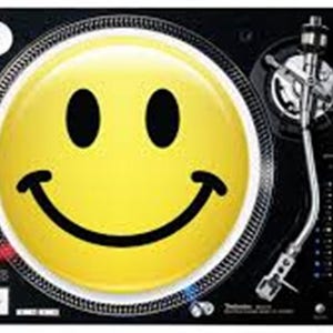 Puede incluir: Un tocadiscos negro con una gran cara sonriente amarilla en el plato. El tocadiscos tiene un brazo de tono plateado y botones de control. La imagen está sobre un fondo negro.