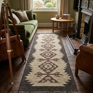 Peut inclure: Un tapis rectangulaire avec un motif géométrique. Le centre est beige, bordé de marron foncé. Des motifs de losanges et de flèches, marron et bordeaux, ornent le tapis. Des franges sont présentes aux deux extrémités.