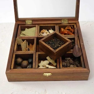 May include: A square wooden spice box with a hinged glass lid and brass hardware. The interior features nine compartments containing various dried spices, including black peppercorns, cinnamon sticks, and bay leaves, with a small wooden spoon.