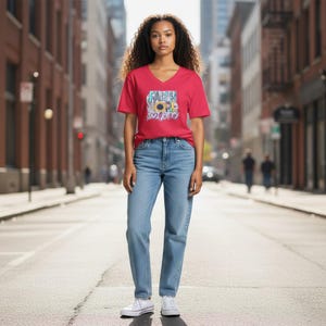 Puede incluir: Una camiseta roja con cuello en V con un diseño gráfico con las palabras "Fe, Amor" y un girasol. La camiseta se combina con vaqueros azul claro y zapatillas blancas. La modelo está de pie en una calle de la ciudad.