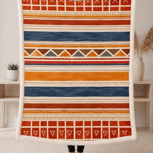 May include: A rectangular throw blanket featuring a white sherpa fleece border and a repeating horizontal pattern of orange, red, and blue stripes, interspersed with geometric shapes including triangles, squares, and thin vertical lines.