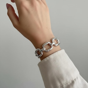 Puede incluir: Un brazalete plateado con eslabones de cadena. El brazalete presenta una serie de eslabones rectangulares interconectados. Los eslabones son lisos y tienen un acabado pulido. El brazalete se lleva en la muñeca y el fondo es de color neutro.