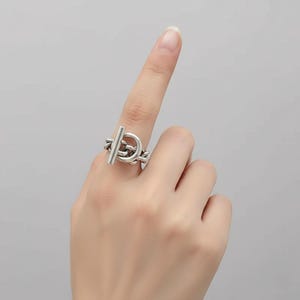May include: A silver-toned ring featuring a toggle clasp design. The ring has a bar that slides through a loop, creating a secure closure. The ring is worn on a finger, showcasing its intricate details and modern aesthetic.