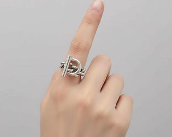 以下が含まれることがあります： トグルクラスプのデザインが特徴のシルバーリング。リングには、ループをスライドするバーがあり、安全な留め具を作成します。リングは指に着用され、その複雑なディテールとモダンな美学を披露しています。