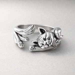 May include: A silver-colored ring featuring a detailed, three-dimensional cat design. The cat is sculpted in a relaxed pose, with its paws and tail forming the ring's band. The cat's face is detailed with closed eyes and a gentle expression.