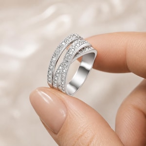May include: A silver-toned ring featuring a crossover design with three distinct bands. The two outer bands are encrusted with small, clear, sparkling stones, while the central band is smooth and polished, creating a layered, multi-strand appearance.