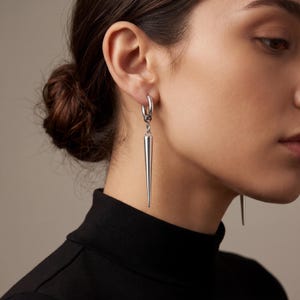 以下が含まれることがあります： 長く尖った円錐形のペンダントが付いた、シルバーのフープピアス。ピアスは揺れており、研磨された反射面を持っています。ジュエリーは人の耳に着用されており、背景には黒のタートルネックがあります。