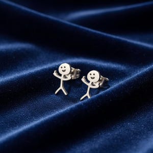 May include: A pair of silver stick figure stud earrings. Each earring features a small, smiling stick figure with arms raised in a joyful pose. The earrings are set against a dark blue, textured background.