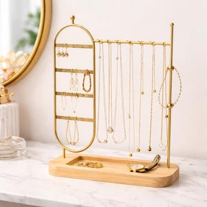 May include: A gold-colored jewelry organizer with a wooden base. The organizer features a vertical earring display and a horizontal bar for necklaces. Several gold necklaces and earrings are displayed on the organizer, along with a watch and rings.