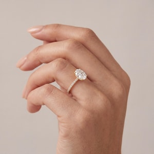 May include: A gold engagement ring featuring an oval-cut diamond. The band is embellished with tiny, glittering stones. The ring is displayed on a hand against a plain backdrop. The ring is a symbol of commitment and love.