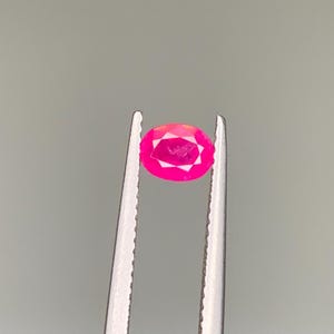 Peut inclure: Un rubis de forme ovale, maintenu par une pince. Le rubis présente une couleur rose intense, avec des facettes captant la lumière. La pierre précieuse est au centre, sur un fond neutre, mettant en valeur sa clarté et sa couleur.
