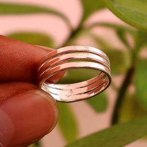 Puede incluir: Un anillo de tono plateado pulido con un diseño de triple banda. El anillo consta de tres bandas metálicas delgadas, paralelas y lisas que están unidas para formar una sola pieza de joyería cohesiva con un acabado metálico reflectante.