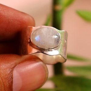 Può includere: Un anello a fascia larga in argento con un cabochon centrale in pietra di luna di forma ovale. La superficie metallica dell'anello presenta una finitura martellata e testurizzata, e la pietra mostra un sottile riflesso blu iridescente.