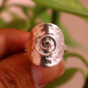 May include: A silver-toned ring featuring a large, circular, hammered metal face. The center of the disc displays a raised, coiled spiral design. The band is thin and metallic, connecting to the back of the textured, reflective circular centerpiece.