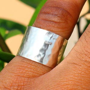 以下が含まれることがあります： 指にはめられた、幅広のハンマー加工されたシルバーリング。 リングはテクスチャ加工された表面とシンプルでモダンなデザインです。 背景には緑の葉とニュートラルな色調の背景があります。