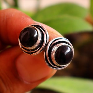 Puede incluir: Un par de pendientes de botón con piedras de ónice negro pulidas y redondas, engastadas en monturas circulares de plata oxidada con detalles de alambre concéntrico. Los pendientes tienen un acabado metálico oscuro y un perfil redondeado.