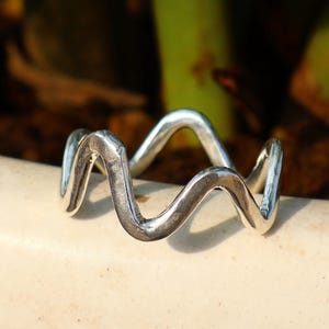 Puede incluir: Un anillo plateado con un diseño ondulado. El anillo presenta un patrón continuo y ondulado, que se asemeja a una ola estilizada. El anillo es una pieza de joyería y está sobre un fondo neutro.