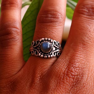 Op de afbeelding: Een zilveren ring met een ovale, blauwe edelsteen gezet in een sierlijk filigraan ontwerp. De ring zit om een vinger, met een groene plant op de achtergrond.