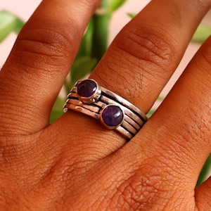 Könnte beinhalten: Ein mehrreihiger Silberring mit zwei runden, polierten violetten Amethyst-Edelsteinen in individuellen Fassungen. Der Ring besteht aus mehreren dünnen, übereinanderliegenden Silberbändern, die eine geschichtete Optik erzeugen.