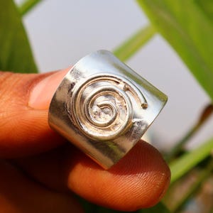 May include: A wide silver band ring featuring a raised spiral design on the front surface. The metal has a polished, reflective finish, and the spiral pattern is centered on the band, creating a distinct texture against the smooth silver surface.