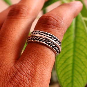 Op de afbeelding: Een zilveren ring met een gevlochten touwontwerp wordt aan een vinger gedragen. De ring heeft een getextureerd uiterlijk met meerdere banden. De achtergrond bevat groene bladeren, wat wijst op een natuurlijke omgeving. De ring is een sieraad.