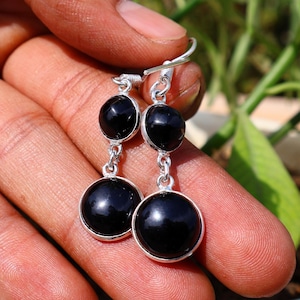 Puede incluir: Pendientes colgantes de plata con tres piedras de ónix negras y redondas. Las piedras están engarzadas en plata y unidas por cadenas de plata. Los pendientes se sostienen en una mano, con un fondo de hojas verdes.
