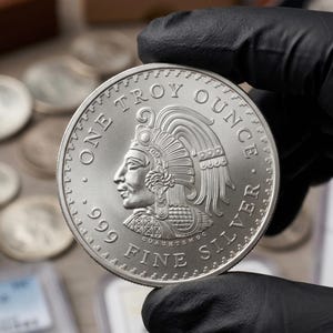 May include: A silver coin featuring a detailed profile of a person wearing a headdress. The coin is marked "ONE TROY OUNCE .999 FINE SILVER" and has a textured edge. The coin is held by a gloved hand.