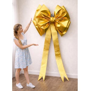 May include: A large, golden bow with long streamers hangs on a wall. The bow is made of a shiny, metallic material. The bow is a decorative element, perfect for events and celebrations.