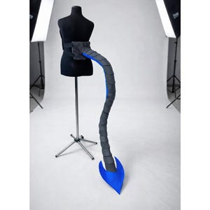 May include: A black mannequin torso with a grey and blue tail prop. The tail is segmented, with a dark grey exterior and blue interior. The prop attaches to a black belt worn around the mannequin's waist.