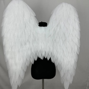 Puede incluir: Grandes alas de ángel blancas con un diseño de plumas en capas. Las alas se exhiben sobre un maniquí negro sobre un fondo blanco. Las alas están hechas de un material texturizado.