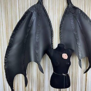 Puede incluir: Alas de disfraz negras con una apariencia texturizada y escamosa, exhibidas en un maniqu&iacute; negro. Las alas tienen puntas puntiagudas y un dise&ntilde;o en capas. Una flor rosa adorna el pecho del maniqu&iacute; y un cintur&oacute;n plateado rodea la cintura.