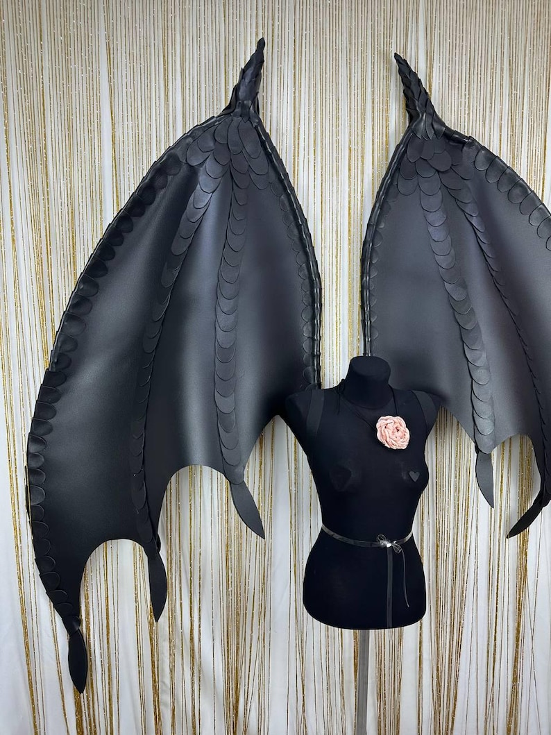 Puede incluir: Alas de disfraz negras con una apariencia texturizada y escamosa, exhibidas en un maniqu&iacute; negro. Las alas tienen puntas puntiagudas y un dise&ntilde;o en capas. Una flor rosa adorna el pecho del maniqu&iacute; y un cintur&oacute;n plateado rodea la cintura.