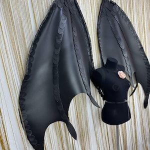 Puede incluir: Alas de drag&oacute;n de cuero sint&eacute;tico negro con un dise&ntilde;o de borde festoneado. Las alas se exhiben en un maniqu&iacute; negro con un acento de flor rosa. Accesorio de disfraz.