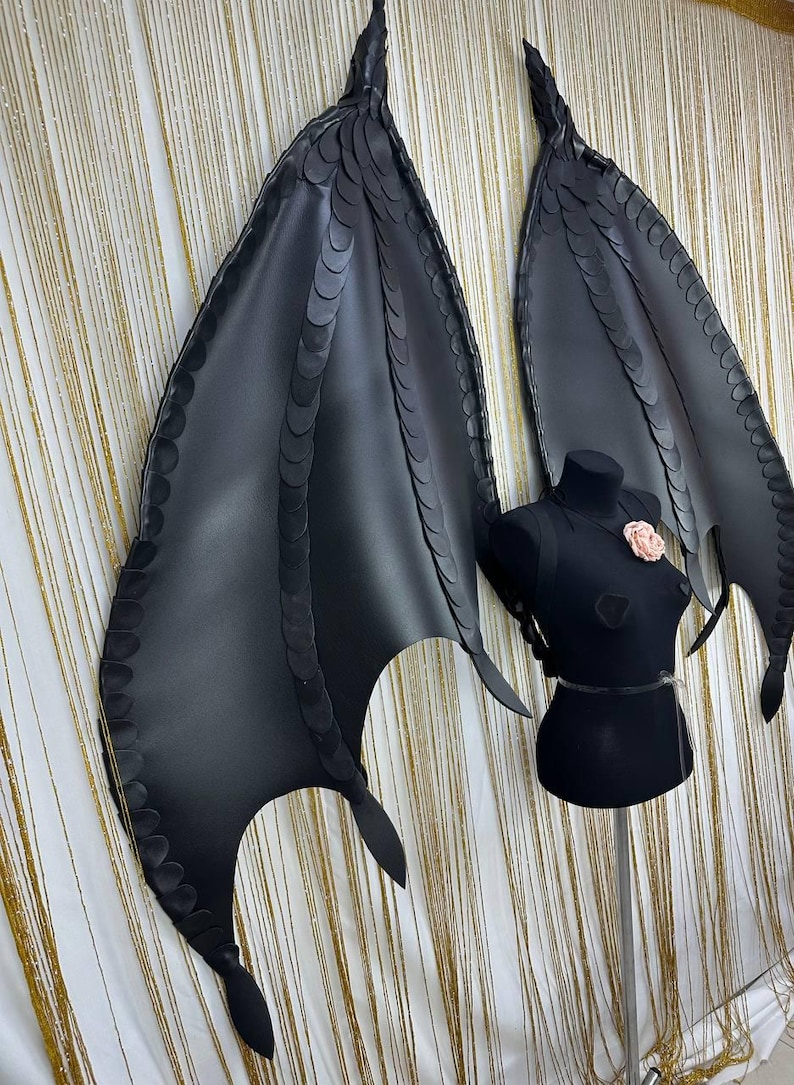 Puede incluir: Alas de drag&oacute;n de cuero sint&eacute;tico negro con un dise&ntilde;o de borde festoneado. Las alas se exhiben en un maniqu&iacute; negro con un acento de flor rosa. Accesorio de disfraz.
