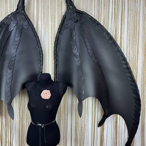 Puede incluir: Alas de drag&oacute;n negras de cuero sint&eacute;tico con textura de escamas y puntas afiladas. Las alas se exhiben en un maniqu&iacute; negro con un adorno floral rosa. Un accesorio de disfraz.