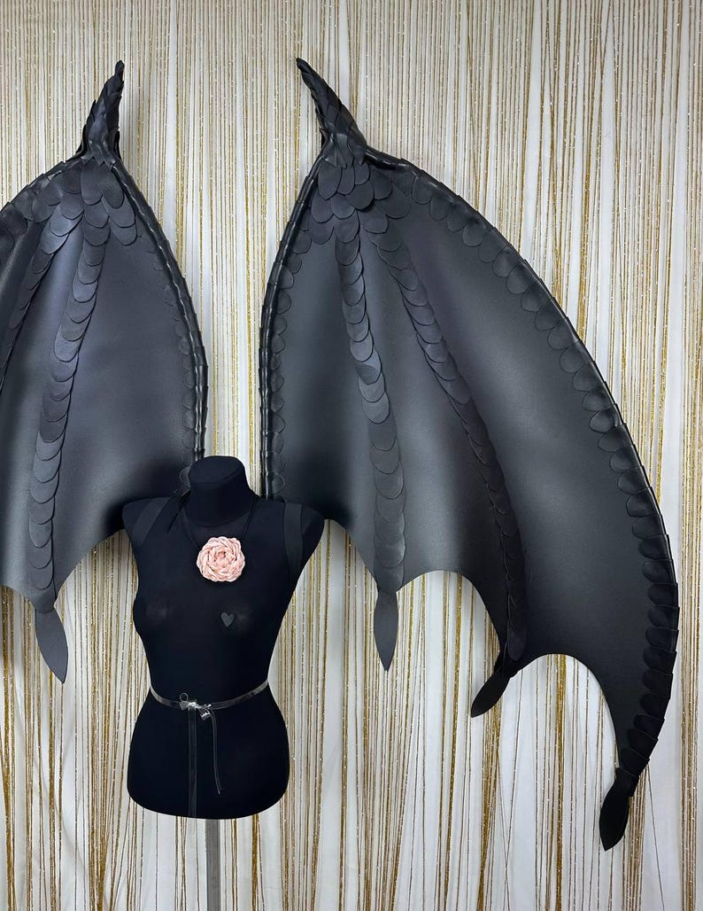 Puede incluir: Alas de drag&oacute;n negras de cuero sint&eacute;tico con textura de escamas y puntas afiladas. Las alas se exhiben en un maniqu&iacute; negro con un adorno floral rosa. Un accesorio de disfraz.
