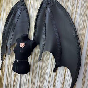 Puede incluir: Grandes alas de disfraz negras con textura escamosa, expuestas sobre un maniqu&iacute; negro. Las alas son detalladas, con forma curva y puntas afiladas. Una flor rosa adorna el pecho del maniqu&iacute;. El fondo es una cortina dorada.