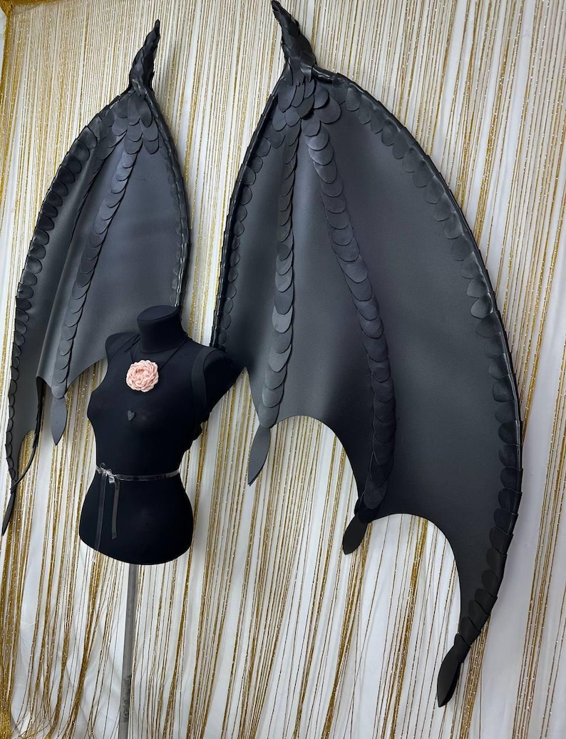 Puede incluir: Grandes alas de disfraz negras con textura escamosa, expuestas sobre un maniqu&iacute; negro. Las alas son detalladas, con forma curva y puntas afiladas. Una flor rosa adorna el pecho del maniqu&iacute;. El fondo es una cortina dorada.