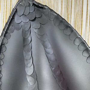 Puede incluir: Un accesorio en forma de ala gris oscuro con un patr&oacute;n texturizado de escamas. El ala tiene una punta y una serie de escamas semicirculares superpuestas a lo largo de los bordes y la columna vertebral. El fondo es una tela a rayas doradas y blancas.
