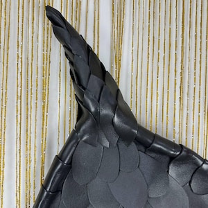 Puede incluir: Bolso triangular de cuero negro con un patr&oacute;n texturizado en forma de escamas. El bolso tiene una parte superior puntiaguda y un asa de cuero trenzado. El fondo es una cortina a rayas doradas y blancas.