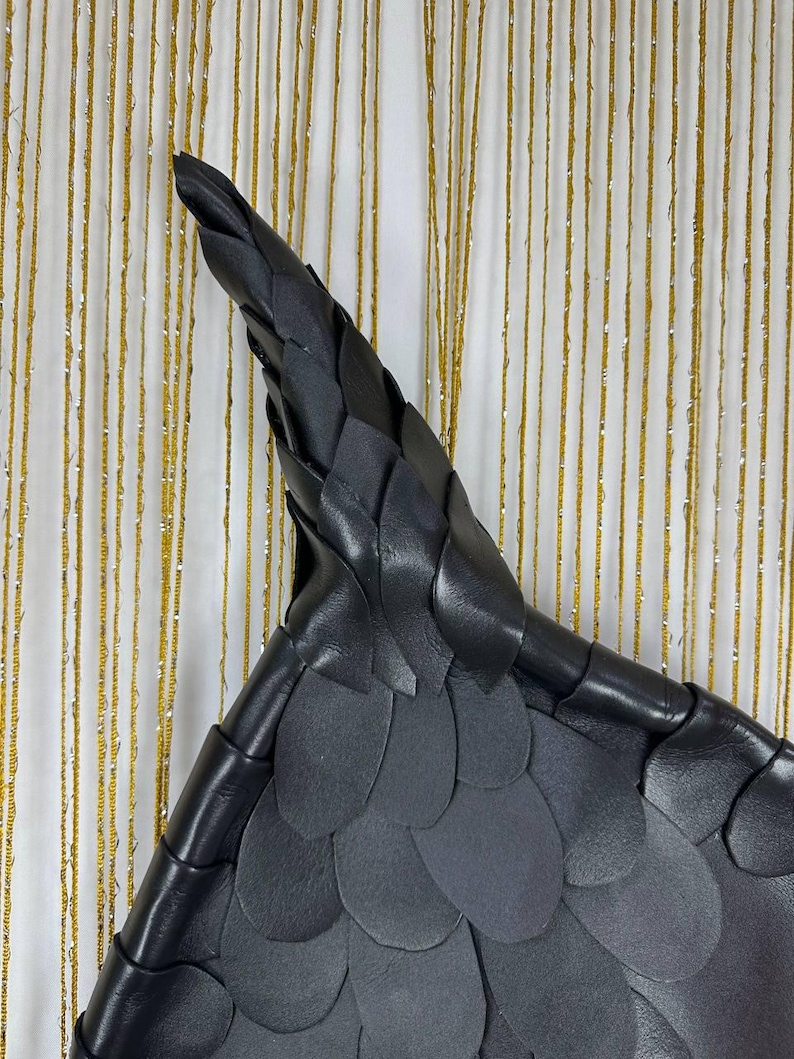 Puede incluir: Bolso triangular de cuero negro con un patr&oacute;n texturizado en forma de escamas. El bolso tiene una parte superior puntiaguda y un asa de cuero trenzado. El fondo es una cortina a rayas doradas y blancas.